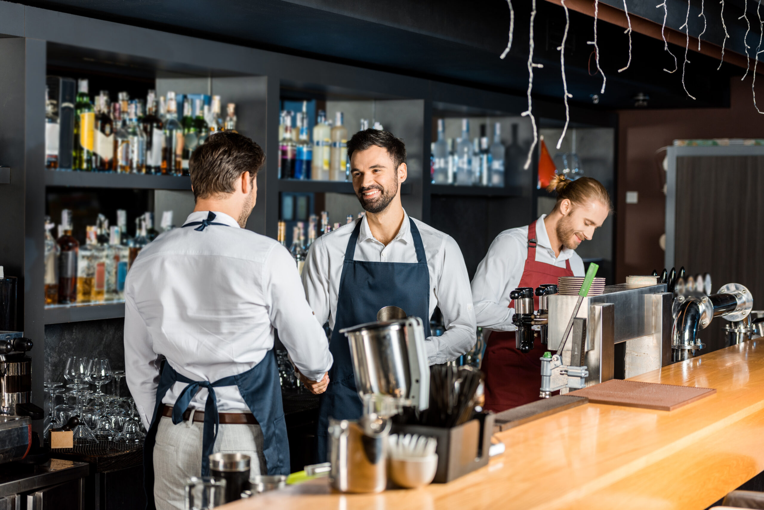 adult barmen in aprons shaking hands near counter